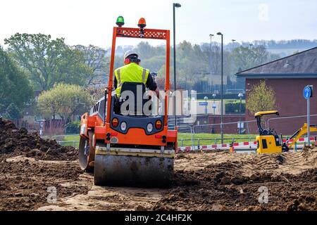 Un travailleur portant un casque de sécurité, un gilet haute visibilité et un casque antibruit entraîne un rouleau de terre sur le chantier Banque D'Images