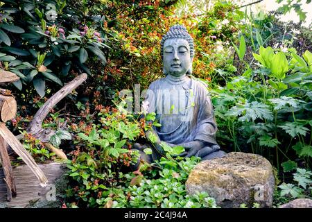 Figurine Bouddha assise médiante, située dans le jardin de feuilles vertes et de petites fleurs avec des rochers et de vieilles branches Banque D'Images