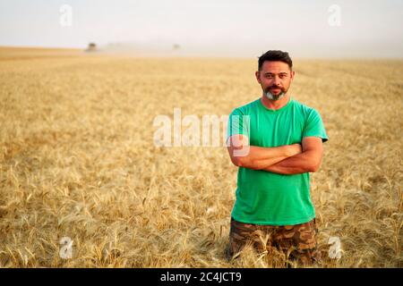 Heureux fermier fièrement debout dans le champ de blé avec les bras croisés sur la poitrine. Agronome portant un uniforme d'entreprise, regardant la caméra sur les terres agricoles. Riche Banque D'Images