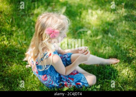 Jeune fille pensive en robe bleue assise sur le sol à l'extérieur l'été ensoleillé jour. Mignon joli petit enfant rêve enfant pensant sur la prairie dehors. AUT Banque D'Images