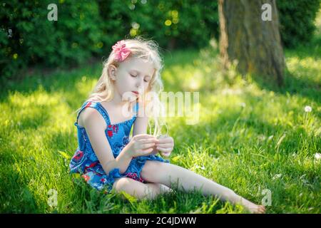 Jeune fille pensive en robe bleue assise sur le sol à l'extérieur l'été ensoleillé jour. Mignon joli petit enfant rêve enfant pensant sur la prairie dehors. AUT Banque D'Images