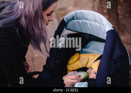 Mère qui allaite bébé fils au biberon dans une poussette à bébé au parc pendant l'automne Banque D'Images