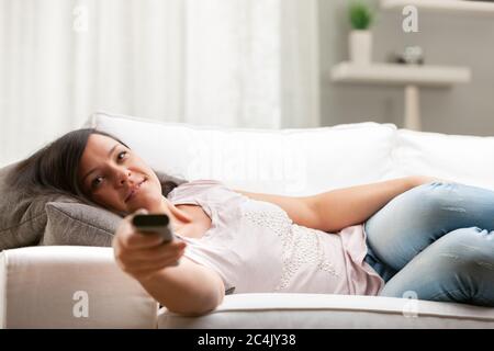 femme souriante aux cheveux longs sur un boush dans son salon changeant de chaîne avec une télécommande Banque D'Images