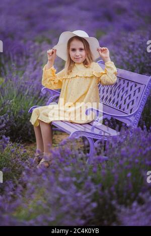 Petite fille souriante en robe jaune et chapeau blanc assis sur un banc violet dans un champ de lavande Banque D'Images