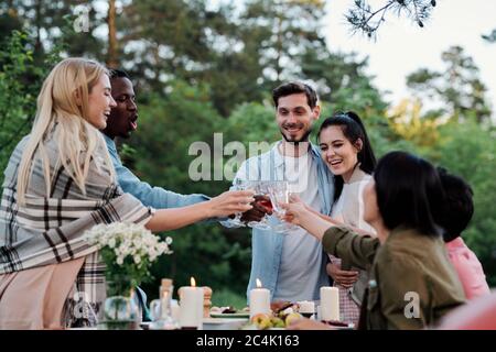 Jeunes amis joyeux de diverses origines ethniques qui se réjouissent avec des verres de vin sur la table servie avec de la nourriture maison et des bougies allumées Banque D'Images