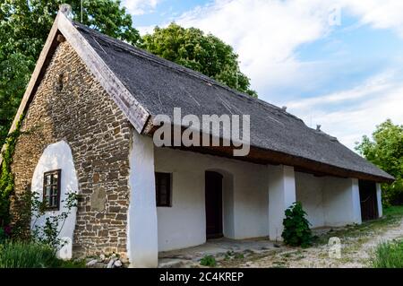 Maison traditionnelle en chaume avec murs blanchis à la chaux et façade en pierre, arbres et plantes dans la cour et ciel bleu au coucher du soleil. Banque D'Images