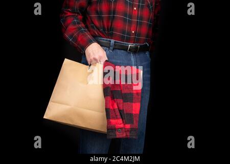 un homme en jean bleu et une chemise à carreaux rouges tenant un sac à provisions en papier avec une autre chemise à carreaux rouges sur fond noir Banque D'Images