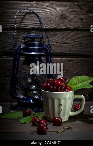 Ancienne lampe à kérosène avec une tasse blanche remplie de cerises mûres sur une table en bois. Encore une vie dans un style rustique. Banque D'Images