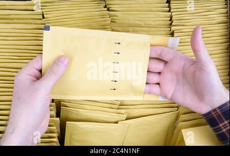 Les mains de l'homme ouvrent une enveloppe à bulles jaunes contre la boîte d'expédition ouverte pleine de pochettes d'expédition rembourrées. Gros plan, vue de dessus. Contrôle qualité de l'auto-scellé Banque D'Images