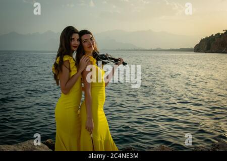 Deux belles filles violinistes dans des robes de concert jaunes jouent des violons électriques avec le coucher du soleil, les montagnes et la mer Méditerranée sur le fond. Banque D'Images