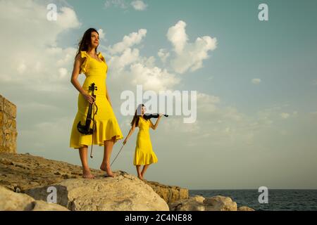 Deux belles sœurs jumelles violinistes dans des robes de concert jaunes jouent des violons électriques sur les rochers au bord de la mer Méditerranée à Antalya, Turquie. Mer Banque D'Images