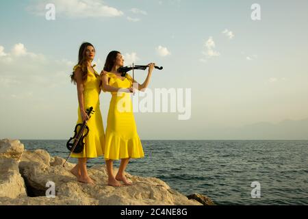 Deux belles sœurs jumelles violinistes dans des robes de concert jaunes jouent des violons électriques sur les rochers au bord de la mer Méditerranée à Antalya, Turquie. PE Banque D'Images