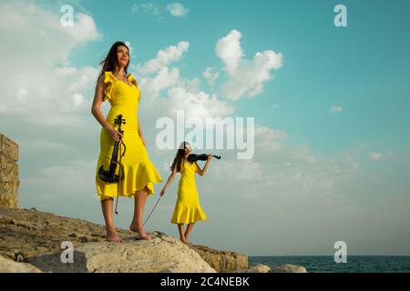 Deux belles sœurs jumelles violinistes dans des robes de concert jaunes jouent des violons électriques sur les rochers au bord de la mer Méditerranée à Antalya, Turquie. Mer Banque D'Images