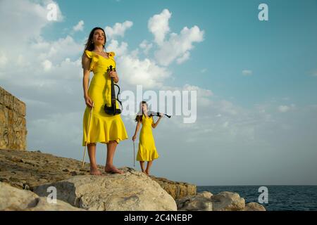 Deux belles sœurs jumelles violinistes dans des robes de concert jaunes jouent des violons électriques sur les rochers au bord de la mer Méditerranée à Antalya, Turquie. Mer Banque D'Images