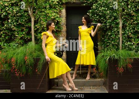 Deux belles sœurs jumelles violinistes dans des robes de concert jaunes posent avec des violons électriques à la porte historique en métal entouré de fleurs en o Banque D'Images