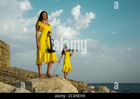 Deux belles sœurs jumelles violinistes dans des robes de concert jaunes jouent des violons électriques sur les rochers au bord de la mer Méditerranée à Antalya, Turquie. Mer Banque D'Images