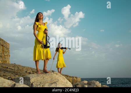 Deux belles sœurs jumelles violinistes dans des robes de concert jaunes jouent des violons électriques sur les rochers au bord de la mer Méditerranée à Antalya, Turquie. Mer Banque D'Images