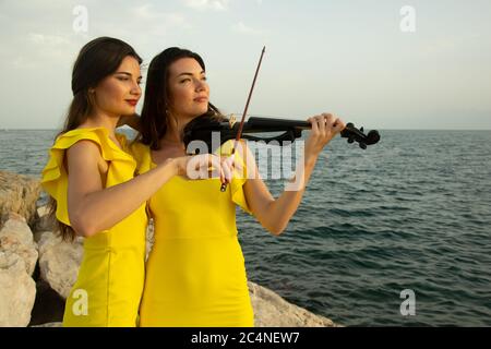 Deux belles filles violinistes dans des robes de concert jaunes jouent des violons électriques avec le coucher du soleil, les montagnes et la mer Méditerranée sur le fond. Banque D'Images