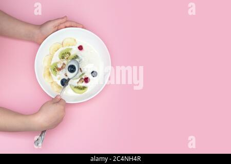 Les mains de l'enfant tiennent un bol et une cuillère de smoothie super-alimentaire. Bol: Yogourt, bleuets, kiwi, banane, noix. Vue de dessus. Banque D'Images