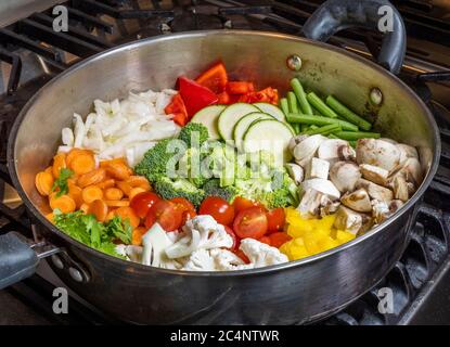 Une casserole ronde en acier sur une cuisinière, pleine de légumes crus coupés en tranches et coupés, prêts à être cuits pour le dîner. Banque D'Images