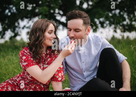 Jeune femme attirée avec un beau homme assis dehors sur une couverture rouge. Femme donner à l'homme fraise. L'homme goûte les baies Banque D'Images