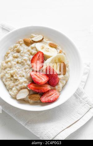 Porridge de flocons d'avoine avec fraises, amandes et banane dans un bol blanc. Petit déjeuner sain avec flocons d'avoine et baies fraîches bio, gros plan. Banque D'Images