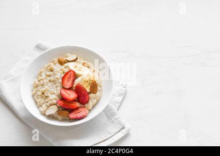 Porridge de flocons d'avoine avec fraises, amandes et banane sur table blanche. Petit déjeuner sain avec flocons d'avoine et baies fraîches bio, espace copie. Banque D'Images