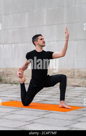 Beau sportif latin homme s'exerçant dans une ville, faisant du yoga, assis dans la variation d'une posture du roi Pigeon à une jambe. Banque D'Images