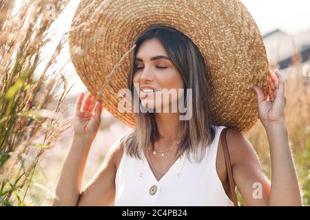 Belle femme blonde touchant son chapeau de paille avec les yeux fermés Banque D'Images