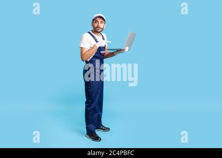 Pleine longueur stupéfait ouvrier en uniforme pointant vers l'ordinateur portable et regardant avec l'expression choquée, surpris par le message sur l'ordinateur, réparation ordre de service. Banque D'Images