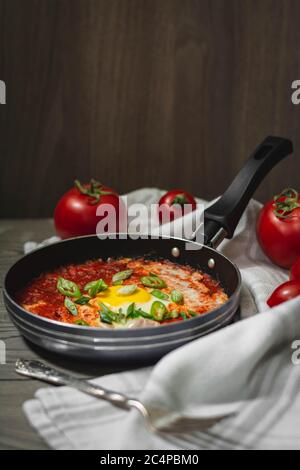 Shakshuka plat traditionnel moyen-oriental avec tomates et œufs dans une petite casserole garnie de gallium et de piment. Tomates en arrière-plan, sombres Banque D'Images