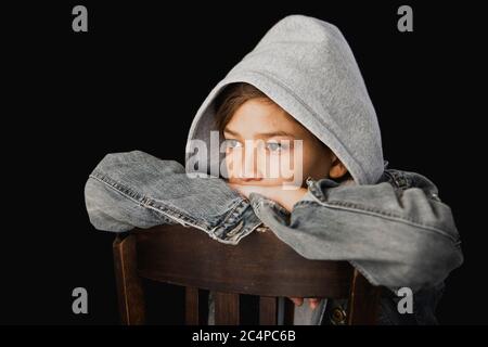 Garçon de 11 ans avec chandail à capuche et veste jean assis sur une chaise en bois sur fond noir Banque D'Images