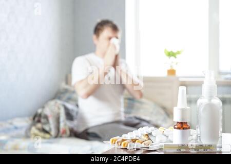 Un malade s'assoit à la maison sur le canapé avec un mouchoir et prend des médicaments. Il y a un thermomètre, des gouttes nasales et des pilules sur la table de nuit Banque D'Images