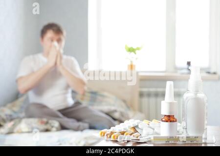 Un malade s'assoit à la maison sur le canapé avec un mouchoir et prend des médicaments. Il y a un thermomètre, des gouttes nasales et des pilules sur la table de nuit Banque D'Images