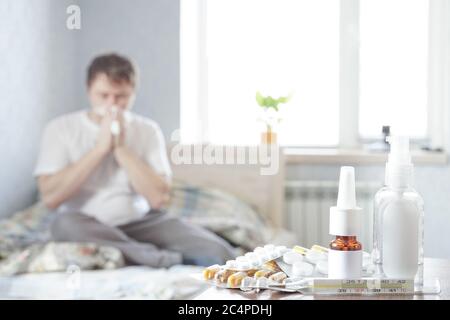 Un malade s'assoit à la maison sur le canapé avec un mouchoir et prend des médicaments. Il y a un thermomètre, des gouttes nasales et des pilules sur la table de nuit Banque D'Images