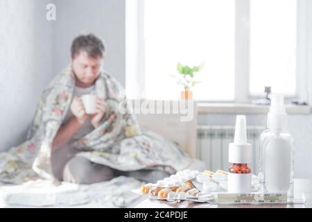 Un malade s'assoit à la maison sur le canapé avec un mouchoir et prend des médicaments. Il y a un thermomètre, des gouttes nasales et des pilules sur la table de nuit Banque D'Images