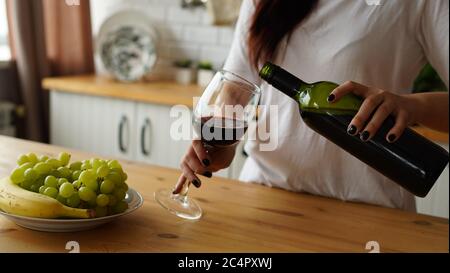 Jeune femme versant du vin rouge dans le verre de la bouteille. Gros plan de la main de la femme remplit le verre transparent avec de l'alcool et le prend. Banque D'Images