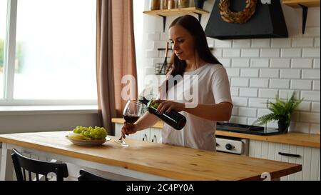 Jeune femme versant du vin rouge dans le verre de la bouteille. Gros plan de la main de la femme remplit le verre transparent avec de l'alcool et le prend. Banque D'Images