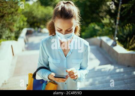 La vie pendant la pandémie du coronavirus. Élégante femme en blouse bleue avec masque médical et sac à main utilisant l'application pour smartphone à l'extérieur dans la rue de la ville. Banque D'Images