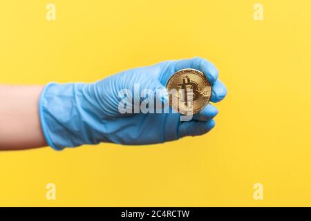 Vue latérale du profil gros plan de la main humaine dans des gants chirurgicaux bleus tenant et montrant le symbole bitcoin en main. Vue en studio, à l'intérieur, isolée sur une bande jaune Banque D'Images