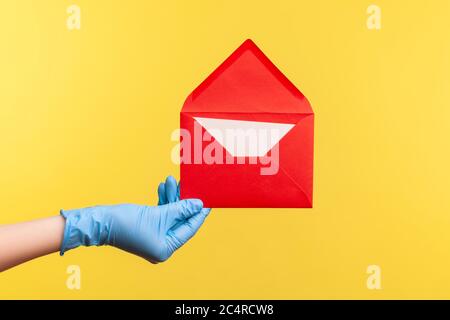 Vue latérale du profil gros plan de la main humaine avec des gants chirurgicaux bleus tenant une enveloppe rouge ouverte. Vue intérieure, studio, isolée sur fond jaune Banque D'Images