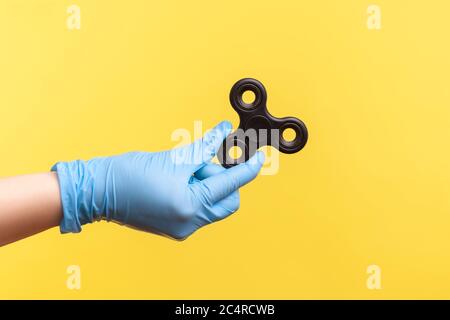 Vue latérale du profil de la main humaine en gants chirurgicaux bleus tenant le spinner de figget noir. Vue en studio, à l'intérieur, isolée sur fond jaune. Banque D'Images