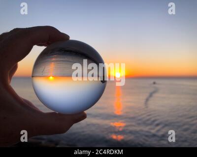 Main de femme tenant une boule de cristal, regardant à travers la mer et le ciel à l'aube. Concept créatif, voyage de rêve. Photographie de réfraction de boule de cristal Banque D'Images