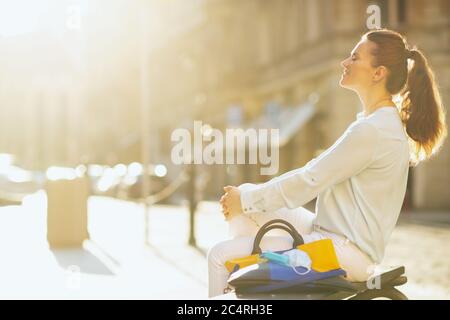 La vie durant la pandémie du coronavirus. Heureuse femme tendance dans un chemisier bleu avec masque médical et sac à main se détendant sur un banc à l'extérieur dans l'ic Banque D'Images