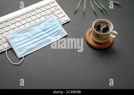 Bureau vue sur le dessus avec clavier, masque médical et tasse de café sur fond noir. Banque D'Images