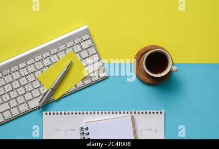 Plat de café, clavier sans fil, bloc-notes, calendrier et papier jaune sur plateau jaune et bleu. Copier l'espace. Banque D'Images