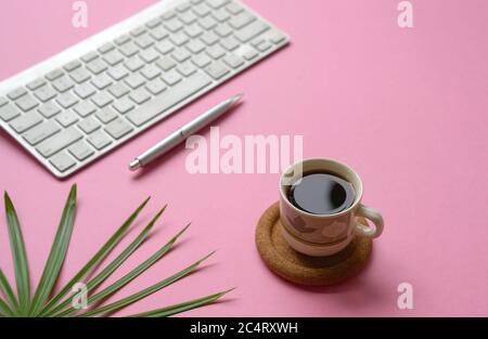 Tasse de café noir sur bureau rose avec clavier, stylo et plante verte. Copier l'espace. Banque D'Images