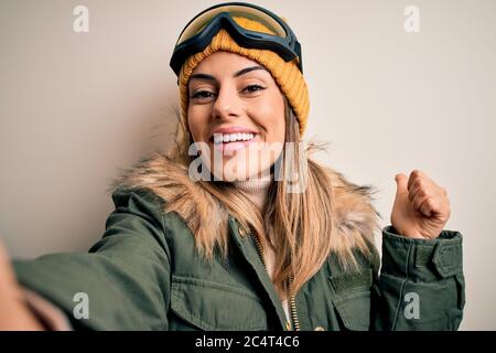 Jeune femme brunette skieur portant des vêtements de neige et des lunettes de ski faire selfie par caméra heureux avec grand sourire faisant signe ok, pouce vers le haut avec les doigts, excellent Banque D'Images