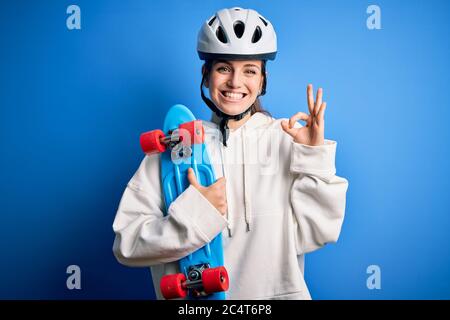Jeune belle redhead skateboarder femme portant un casque de sécurité tenant skate faisant signe ok avec les doigts, excellent symbole Banque D'Images