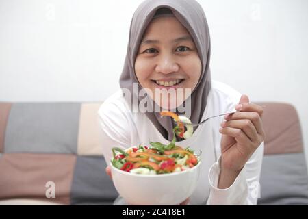 Femme asiatique musulmane portant hijab manger une nourriture saine, salade de légumes verts, après l'exercice à la maison, garder en bonne santé et en forme pendant le nouveau mode de vie normal co Banque D'Images